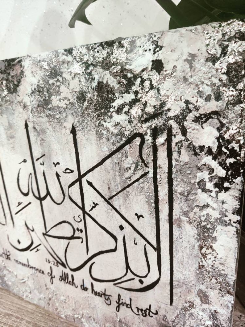 May include: Black and white calligraphy art on a textured canvas background. The Arabic script reads "In the remembrance of Allah do hearts find rest".