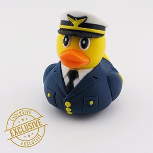 May include: A yellow rubber duck wearing a navy blue pilot uniform, complete with a white hat and black tie. The duck has an orange beak and black eyes. A gold "EXCLUSIVE" stamp is in the lower left corner.
