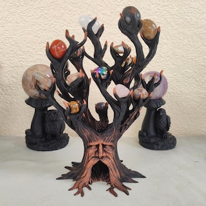 May include: A dark brown tree-shaped sculpture with a carved face at the base and multiple branches holding various polished stones in shades of red, white, pink, and iridescent colors. Two black owl-shaped stands are positioned behind the tree.