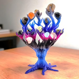 May include: A tree-shaped sculpture with a gradient finish transitioning from black at the base to blue and magenta at the branches. The branches feature multiple prongs holding various polished, multi-colored smooth stones and crystals.