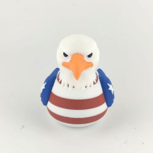 May include: A bald eagle figurine with a white head, orange beak, and a stern expression. The body has red and white stripes, with blue wings featuring white stars, representing the American flag.