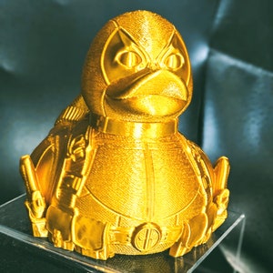 以下が含まれることがあります： 金色の3Dプリントされたデッドプールのフィギュア。スーパーヒーローのデッドプールがアヒルの形をしています。フィギュアは、黒いアイマスクと黒いベルトで詳細に描かれています。