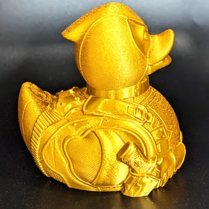 以下が含まれることがあります： スーパーヒーローのコスチュームのような詳細なデザインが施された金色のゴム製のアヒル。アヒルは表面がざらざらしていて、小さな銃のアクセサリーが付いています。