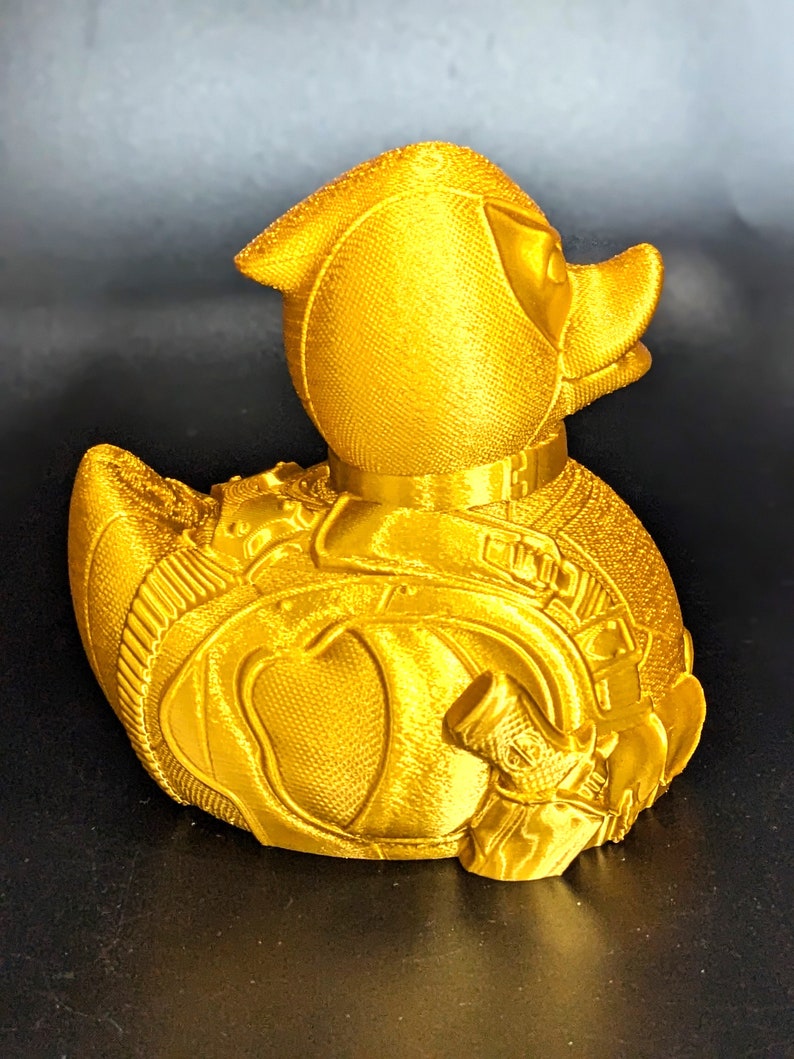 以下が含まれることがあります： スーパーヒーローのコスチュームのような詳細なデザインが施された金色のゴム製のアヒル。アヒルは表面がざらざらしていて、小さな銃のアクセサリーが付いています。