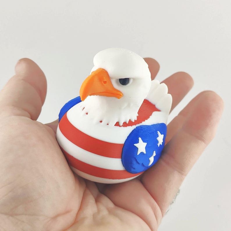 May include: A 3D-printed bald eagle figurine. The eagle has a white head, an orange beak, and a body decorated with red and white stripes and blue sections with white stars, resembling the American flag.