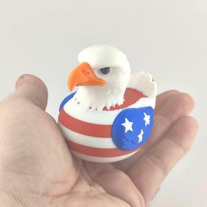 May include: A novelty rubber duck in the shape of a bald eagle, featuring a white head, orange beak, and a red, white, and blue design with stars. A quirky and patriotic item, perfect for collectors or as a gift.