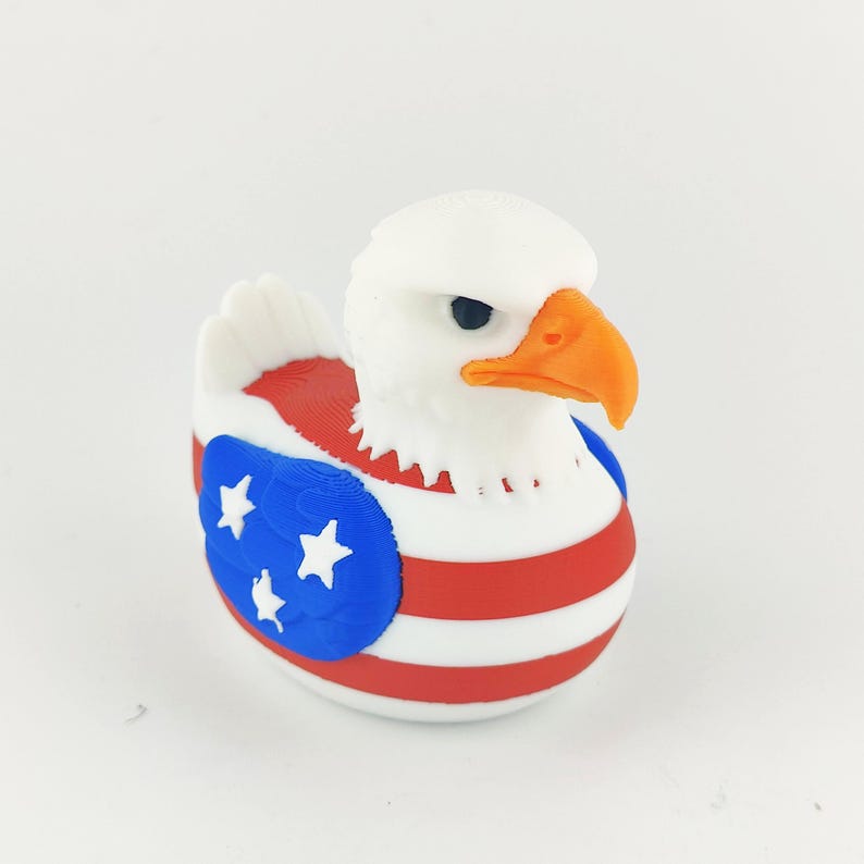 May include: A 3D-printed eagle ornament with a white head and an orange beak. The body features red and white stripes, and a blue section with white stars, mirroring the American flag. The eagle has black eyes.