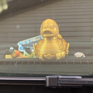 以下が含まれることがあります： デッドプールのデザインが施された金色のゴム製のオモチャの鴨。鴨は他のゴム製の鴨と一緒に車のダッシュボードの上に置かれています。