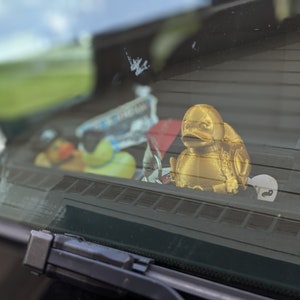 以下が含まれることがあります： 車のダッシュボードに置かれた、金色のゴム製のオモチャの鴨。鴨の背中には、細かい模様が施されています。鴨の周りには、青と白のステッカーなど、他のアイテムが置かれています。