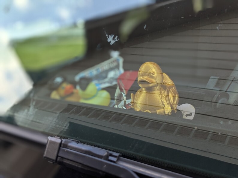 以下が含まれることがあります： 車のダッシュボードに置かれた、金色のゴム製のオモチャの鴨。鴨の背中には、細かい模様が施されています。鴨の周りには、青と白のステッカーなど、他のアイテムが置かれています。