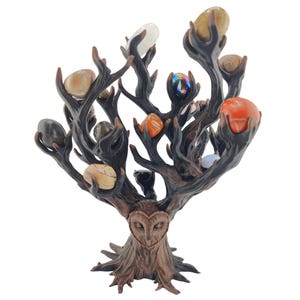 May include: A dark brown, tree-shaped sculpture featuring a sculpted owl face at the base and multiple branches that hold various smooth, polished, multi-colored stones, including orange, white, grey, and iridescent hues, arranged in a cluster.