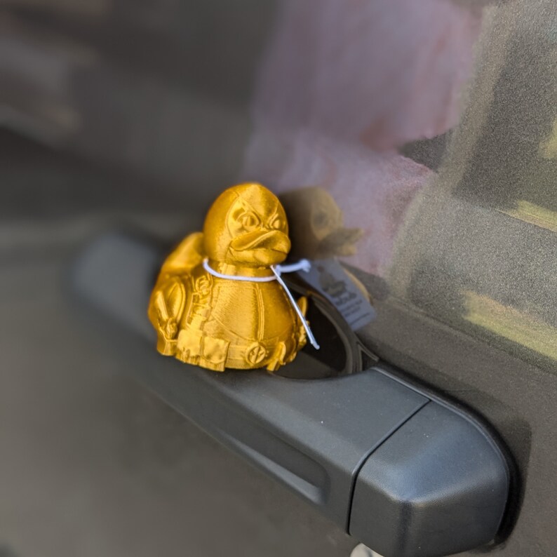 以下が含まれることがあります： 金色の3Dプリントされたゴム製のオモチャの鴨が、黒い車のドアハンドルの上に置かれています。鴨は細部まで丁寧に作られています。