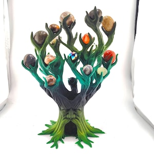 May include: A dark green and black tree-shaped sculpture with a face carved into the trunk. The branches feature multiple pronged holders, each supporting a small, smooth, multi-colored spherical stone in various shades of brown, orange, and red.