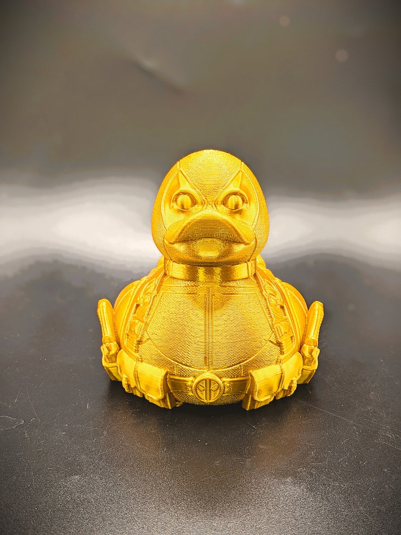 以下が含まれることがあります： デッドプールデザインの金色のゴム製アヒル。アヒルは、黒い目の赤いマスクと、デッドプールのロゴが付いた黒いベルトをしています。