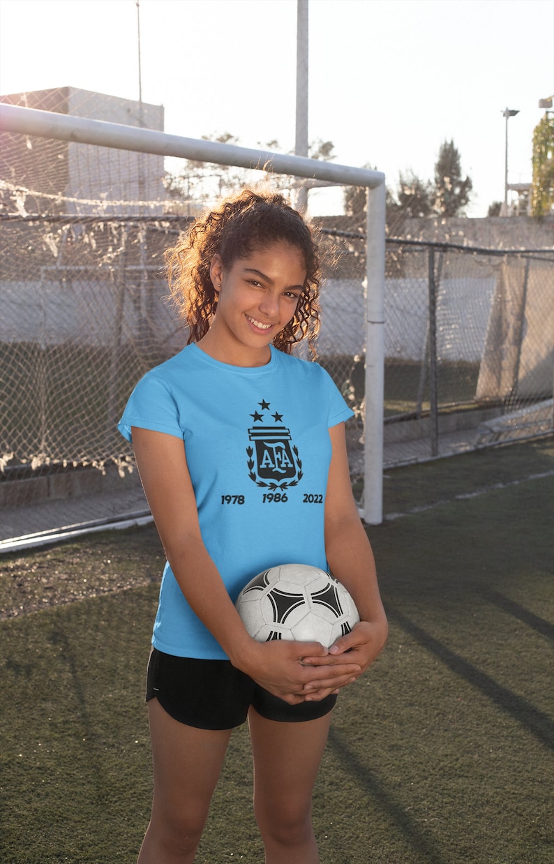 Pu&ograve; includere: Maglietta azzurra con un emblema calcistico nero e gli anni 1978, 1986 e 2022. La persona tiene in mano un pallone da calcio bianco e nero. Lo sfondo mostra un campo da calcio e una porta.