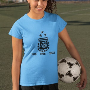 Pu&ograve; includere: Maglietta azzurra con grafica nera di uno scudo, stelle e le lettere "AFA", con gli anni 1978, 1986 e 2022. Un pallone da calcio &egrave; tenuto in mano dalla persona.