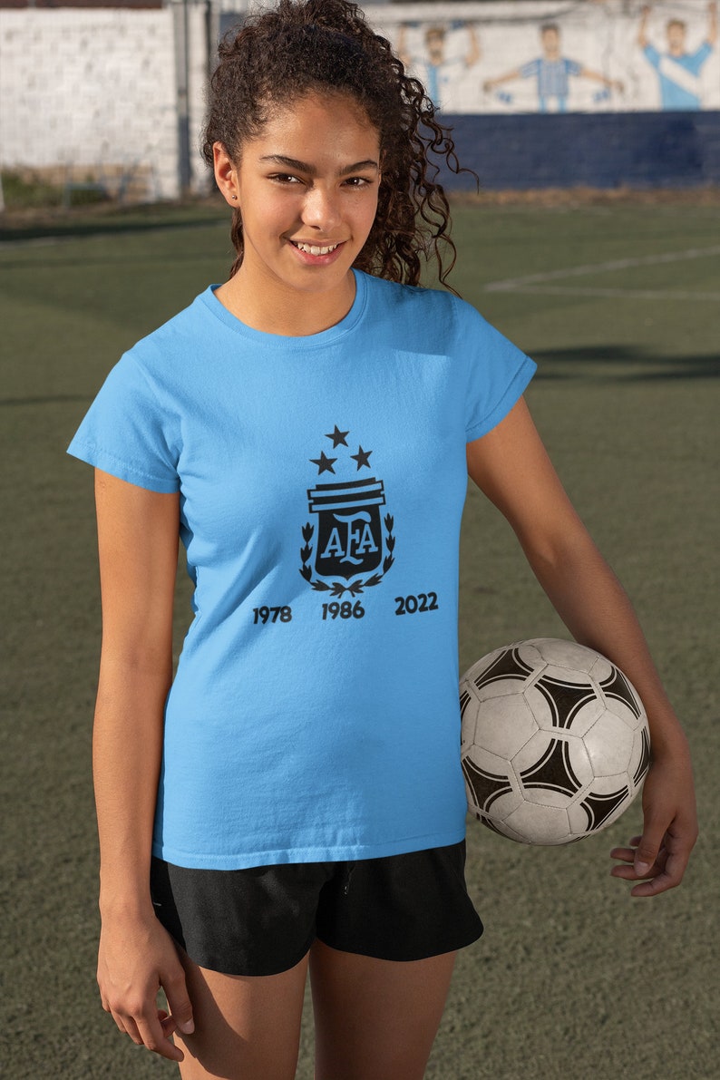 Pu&ograve; includere: Maglietta azzurra con grafica nera di uno scudo, stelle e le lettere "AFA", con gli anni 1978, 1986 e 2022. Un pallone da calcio &egrave; tenuto in mano dalla persona.