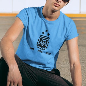 Pu&ograve; includere: Maglietta azzurra con un emblema nero con le lettere "AFA" e tre stelle. La maglietta mostra anche gli anni 1978, 1986 e 2022. Indossata da una persona con berretto grigio e occhiali da sole.