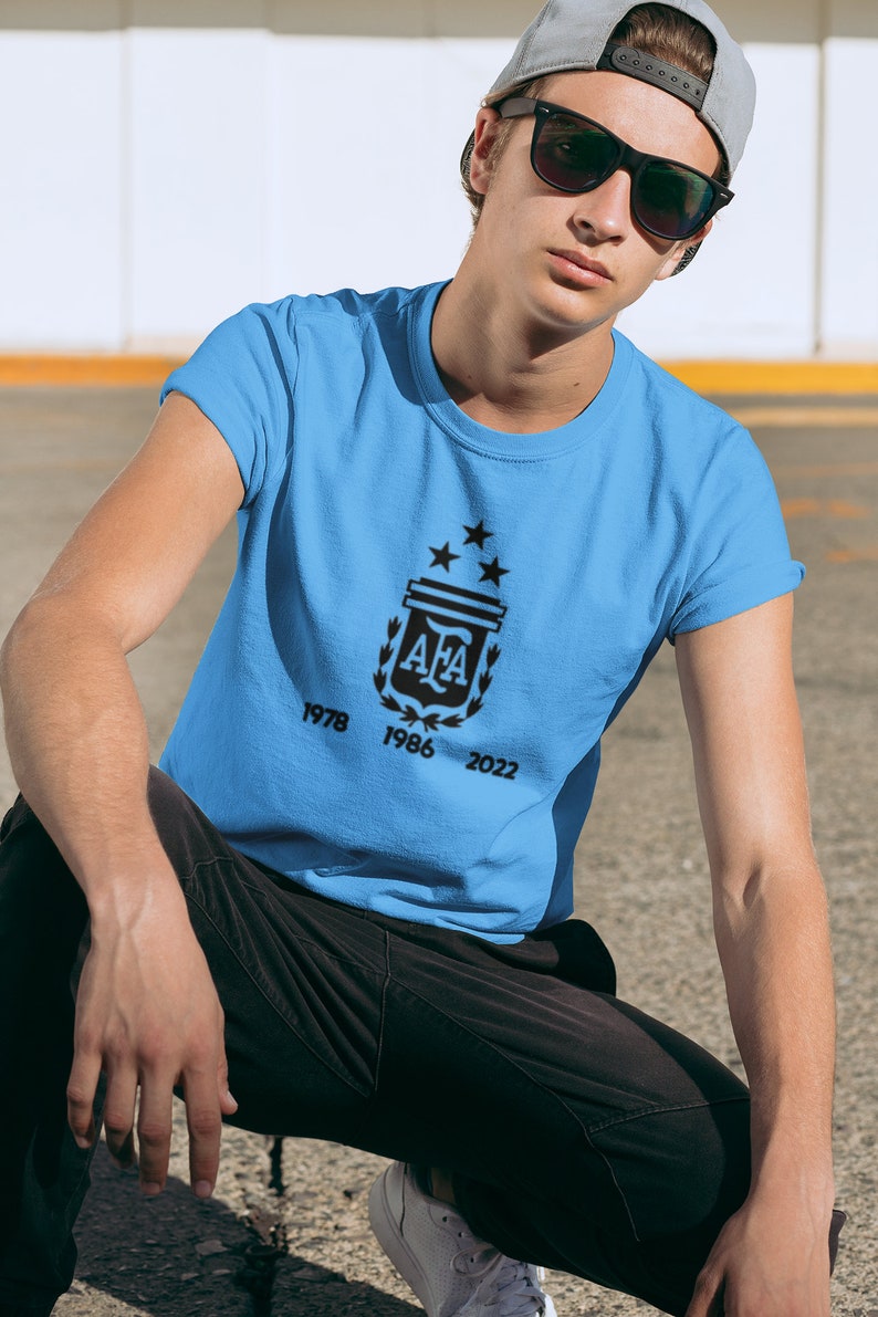 Pu&ograve; includere: Maglietta azzurra con un emblema nero con le lettere "AFA" e tre stelle. La maglietta mostra anche gli anni 1978, 1986 e 2022. Indossata da una persona con berretto grigio e occhiali da sole.