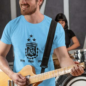 Pu&ograve; includere: Maglietta azzurra con grafica nera del logo dell'Associazione calcistica argentina, tre stelle e gli anni 1978, 1986 e 2022. Una persona tiene in mano una chitarra. &Egrave; visibile una tracolla nera per la chitarra.