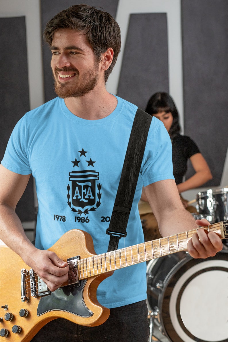 Pu&ograve; includere: Maglietta azzurra con grafica nera del logo dell'Associazione calcistica argentina, tre stelle e gli anni 1978, 1986 e 2022. Una persona tiene in mano una chitarra. &Egrave; visibile una tracolla nera per la chitarra.