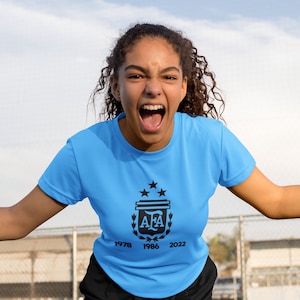 Pu&ograve; includere: Una maglietta azzurra con il logo AFA e gli anni 1978, 1986 e 2022 stampati sotto. La maglietta &egrave; indossata da una persona con capelli ricci scuri, su uno sfondo di cielo.