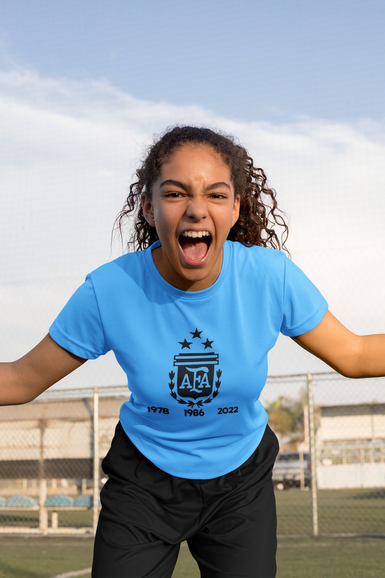 Pu&ograve; includere: Una maglietta azzurra con il logo AFA e gli anni 1978, 1986 e 2022 stampati sotto. La maglietta &egrave; indossata da una persona con capelli ricci scuri, su uno sfondo di cielo.