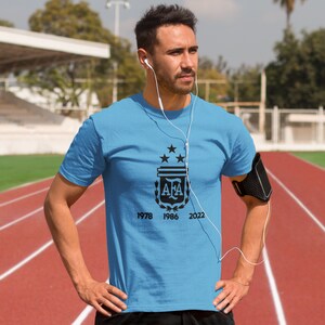 Pu&ograve; includere: Una maglietta azzurra con il logo AFA, tre stelle e gli anni 1978, 1986 e 2022 in nero. La persona indossa la maglietta su una pista di atletica rossa e indossa le cuffie.