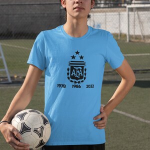 Pu&ograve; includere: Maglietta azzurra con grafica nera di un stemma, tre stelle e le lettere "AFA". Sotto lo stemma ci sono i numeri 1978, 1986 e 2022. Un pallone da calcio &egrave; tenuto in mano.