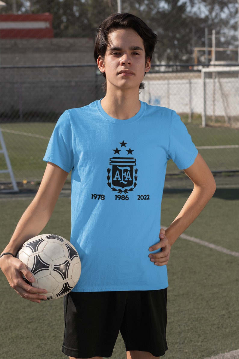 Pu&ograve; includere: Maglietta azzurra con grafica nera di un stemma, tre stelle e le lettere "AFA". Sotto lo stemma ci sono i numeri 1978, 1986 e 2022. Un pallone da calcio &egrave; tenuto in mano.