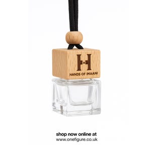 Può includere: Un profumatore per auto in vetro trasparente con una parte superiore in legno e un cordino nero. La parte superiore in legno è incisa con la scritta "HANDS OF IMAANI" e un logo "H" stilizzato. Il profumatore è appeso su sfondo bianco.