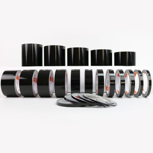Könnte beinhalten: Eine Auswahl an schwarzen Klebebandrollen in verschiedenen Breiten, angeordnet auf weißem Untergrund. Das Klebeband hat eine glänzende Oberfläche, wobei einige Rollen aufrecht stehen und andere flach liegen. Das Bild zeigt verschiedene Größen von Klebeband.