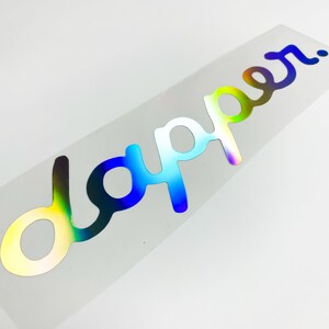 Könnte beinhalten: Ein holografischer Aufkleber mit dem Wort "dapper" in kursivschrift.