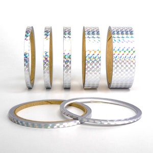 Könnte beinhalten: Ein Satz mit sechs Rollen irisierenden reflektierenden Klebebands in verschiedenen Breiten. Das Klebeband ist silberfarben mit einem holografischen Finish.