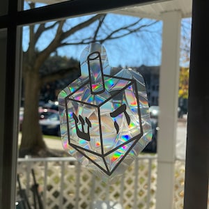 May include: A holographic sticker of a dreidel, a traditional four-sided spinning top used during Hanukkah. The dreidel is decorated with Hebrew letters and a star of David.