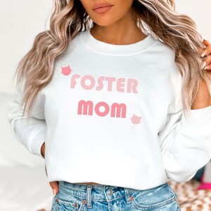 Könnte beinhalten: Weißer Pullover mit dem Text "FOSTER MOM" in rosa Buchstaben und zwei rosa Katzen-Silhouetten.