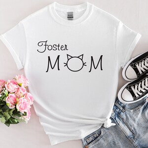 Könnte beinhalten: Weißes T-Shirt mit dem schwarzen Schriftzug "Foster Mom" und einer Katzenkopf-Grafik. Das Shirt wird mit Jeansshorts und schwarzen Converse-Sneakers kombiniert. Ein Strauß rosa Rosen liegt in der Nähe des Shirts.