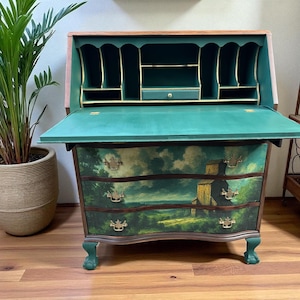 Puede incluir: Un escritorio secretario vintage con una superficie de escritura abatible. El escritorio está pintado en tonos verdes y presenta una pintura de paisaje en los cajones. El escritorio tiene herrajes de latón ornamentados y patas de garra. El escritorio abierto revela estantes y un cajón pequeño.