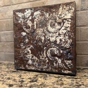 May include: Square, rustic metal wall art with a distressed finish. The artwork features a repeating pattern of swirling designs in shades of brown, white, and hints of blue. The piece has a vintage, antique aesthetic.