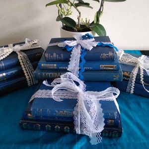 May include: A stack of blue hardback books wrapped with white lace ribbon. The books are arranged in a pyramid shape on a teal blue surface.