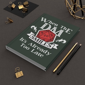 May include: A dark green journal with the text "When The DM Smiles It's Already Too Late" and a red twenty-sided die graphic. The journal is surrounded by gold and black office supplies.
