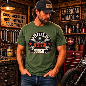 May include: An olive green crew neck t-shirt featuring a graphic print of a piston and two crossed wrenches with the text "BUILT NOT BOUGHT" in a circular emblem. The design includes two red stars and a dark blue background within the circular logo.