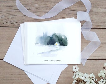 以下が含まれることがあります： 雪景色と小さな家の水彩画が描かれた、空白の白いクリスマスカードのセット。窓は点灯しており、カードには「メリークリスマス！」の文字が印刷されています。