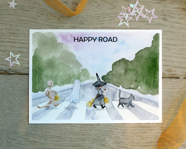 May include: A watercolor painting of a Halloween scene with a ghost, a witch, a cat, and a mummy walking across a crosswalk. The text "HAPPY ROAD" is written at the top of the painting.