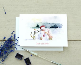 Pode incluir: Cartão de Natal em aquarela com um boneco de neve com chapéu e cachecol vermelhos, um gato, um coelho e o texto "Feliz Natal!"