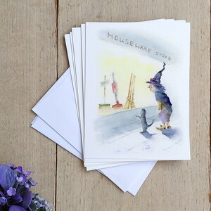 May include: A watercolor illustration of a witch and a black cat standing in front of a store called "Houseware Store". The witch is wearing a purple and black robe and a pointed hat. The cat is looking up at the witch.