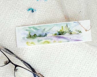 Peut inclure: Marque-page rectangulaire à l'aquarelle représentant un paysage avec des arbres verts, des champs violets et un ciel bleu. Une chaîne dorée est attachée. Le marque-page est sur une surface blanche texturée.