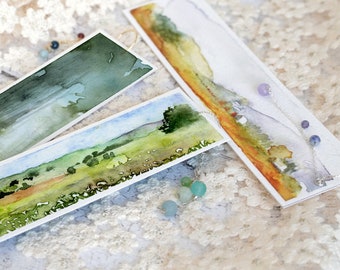 Peut inclure: Trois marque-pages aquarelle avec des scènes de paysages différentes. Les marque-pages sont sur un fond de dentelle blanche avec une chaîne argentée et des perles bleues et violettes.