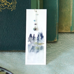 May include: A watercolour bookmark featuring a landscape scene with trees and a lake. The bookmark has a silver chain with beads hanging from the top.