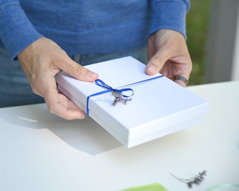 K&ouml;nnte beinhalten: Eine wei&szlig;e Geschenkbox mit einem blauen Band und einem Lavendelzweig oben drauf.